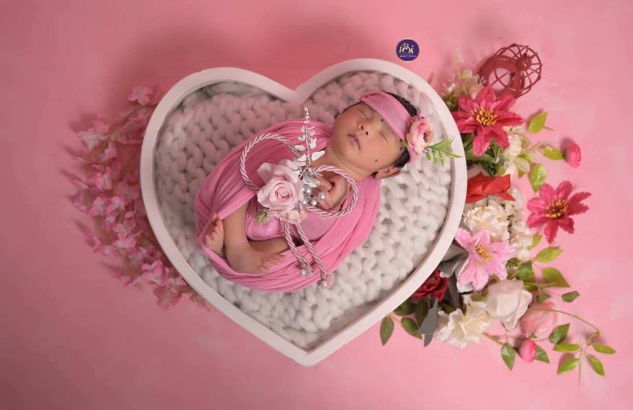 Sleeping newborn baby wrapped in pink fabric inside a heart-shaped basket with floral styling